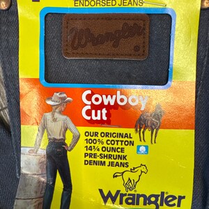 May include: Close-up of Wrangler Cowboy Cut denim jeans. The jeans have a brown leather patch with the Wrangler logo. The tag reads "Our Original 100% Cotton 14 3/4 ounce Pre-shrunk Denim Jeans". The tag also features a drawing of a cowboy and horse.