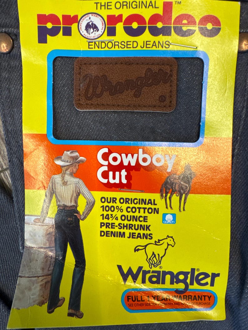 May include: Close-up of Wrangler Cowboy Cut denim jeans. The jeans have a brown leather patch with the Wrangler logo. The tag reads "Our Original 100% Cotton 14 3/4 ounce Pre-shrunk Denim Jeans". The tag also features a drawing of a cowboy and horse.