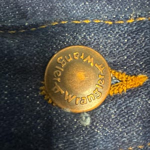 K&ouml;nnte beinhalten: Nahaufnahme eines kupferfarbenen Knopfes mit dem Wort "Wrangler" am Rand. Der Knopf ist an einem dunkelblauen Denimstoff mit gelben N&auml;hten befestigt.