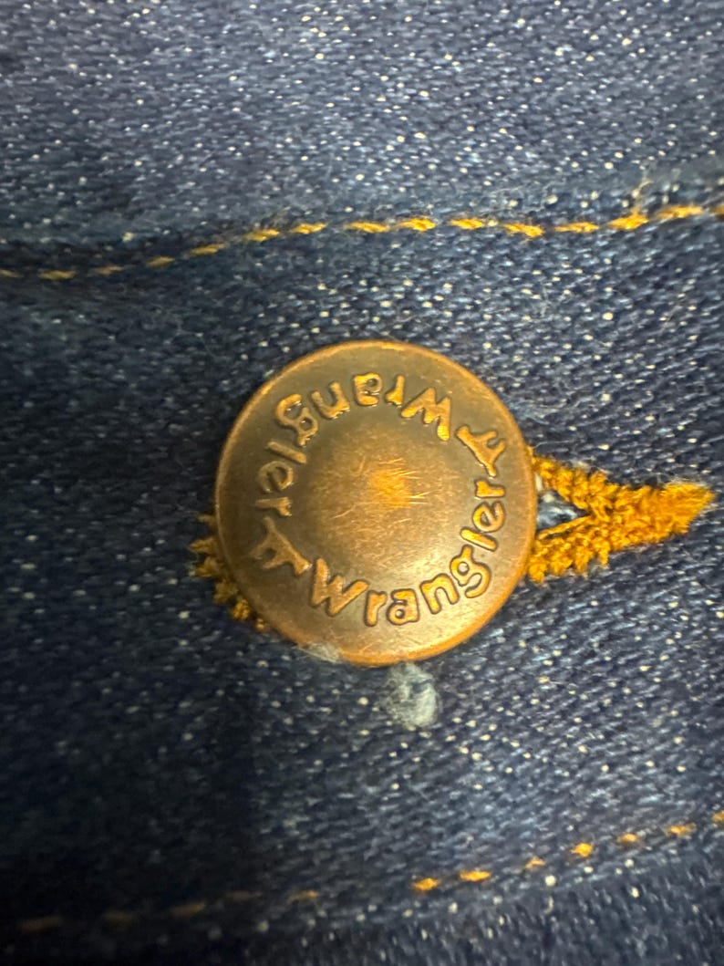 K&ouml;nnte beinhalten: Nahaufnahme eines kupferfarbenen Knopfes mit dem Wort "Wrangler" am Rand. Der Knopf ist an einem dunkelblauen Denimstoff mit gelben N&auml;hten befestigt.