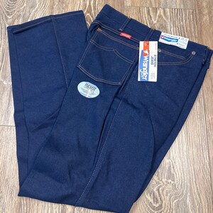 May include: Dark blue Wrangler Flex Fit jeans with a tag. The jeans have a classic five-pocket design with orange stitching. The tag reads "Wrangler Stretch Jeans". The jeans are folded on a wooden surface.