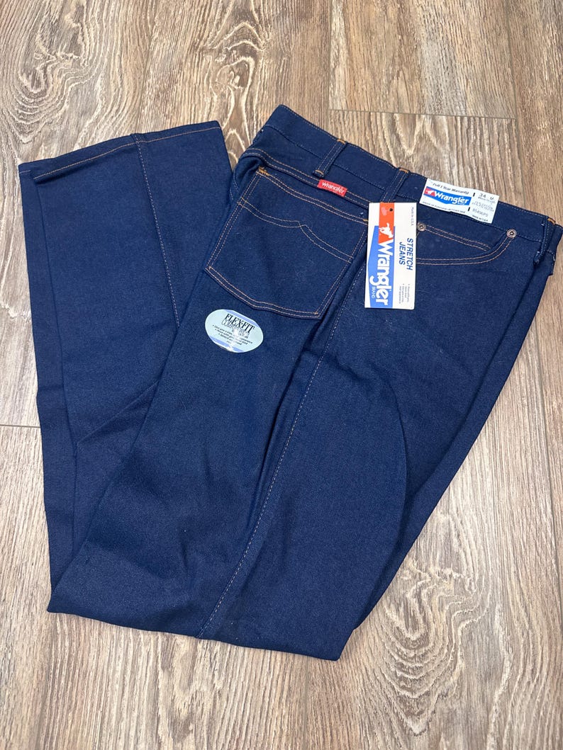 May include: Dark blue Wrangler Flex Fit jeans with a tag. The jeans have a classic five-pocket design with orange stitching. The tag reads "Wrangler Stretch Jeans". The jeans are folded on a wooden surface.