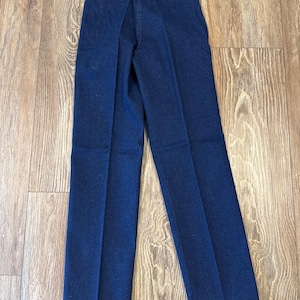 May include: Dark blue denim pants with a straight leg design. The pants have a classic five-pocket style and a Wrangler tag attached. The pants are displayed on a light brown wooden surface.