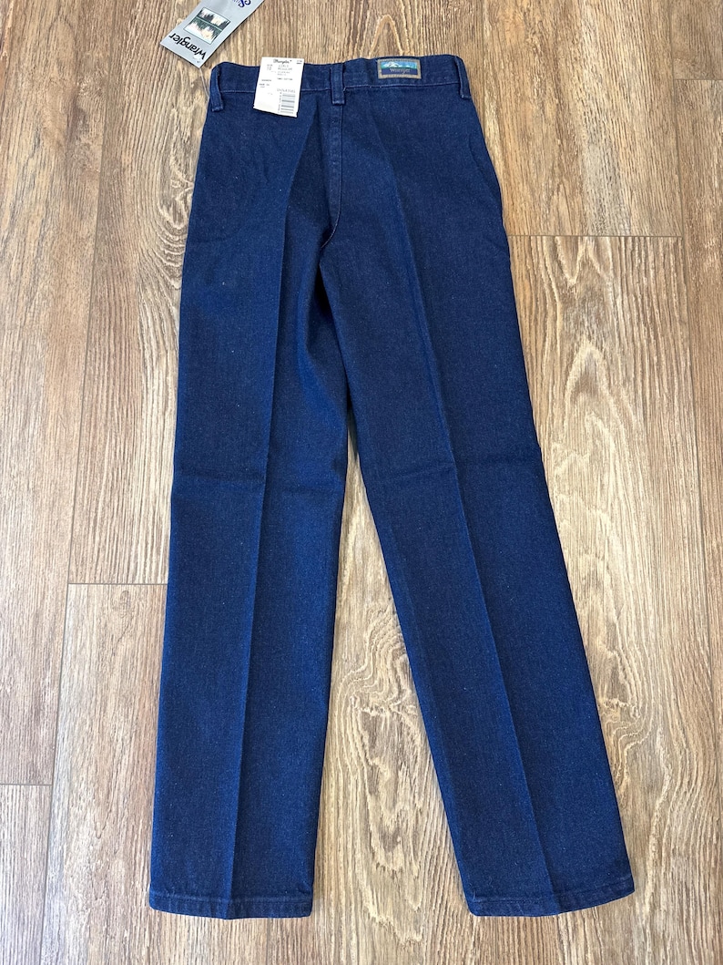 May include: Dark blue denim pants with a straight leg design. The pants have a classic five-pocket style and a Wrangler tag attached. The pants are displayed on a light brown wooden surface.