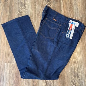 May include: A pair of dark blue denim jeans with a brown leather patch that says "Wrangler" on the back pocket. The jeans have a straight leg and a classic fit.
