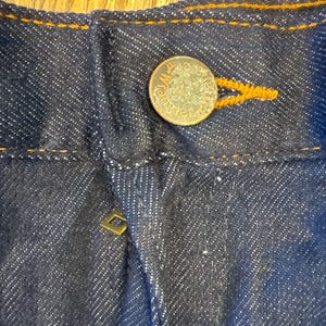 Peut inclure: Gros plan sur la ceinture d'un jean bleu fonc&eacute; avec un bouton en cuivre et un petit rivet dor&eacute;. Les coutures sont de couleur orange contrast&eacute;e.