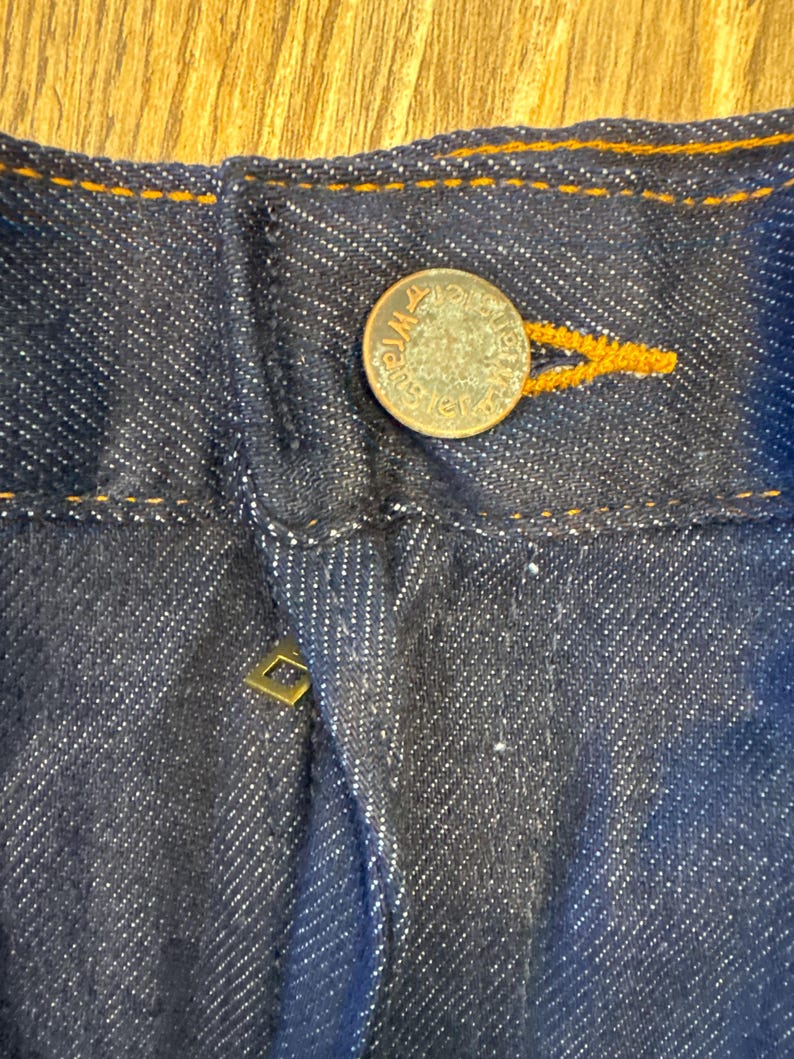 Peut inclure: Gros plan sur la ceinture d'un jean bleu fonc&eacute; avec un bouton en cuivre et un petit rivet dor&eacute;. Les coutures sont de couleur orange contrast&eacute;e.