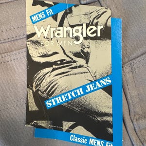 May include: Close-up of light gray Wrangler jeans with a tag. The tag is blue and white with the words "MENS Fit", "Wrangler FOR MEN", and "STRETCH JEANS". The tag also says "Classic MENS Fit".