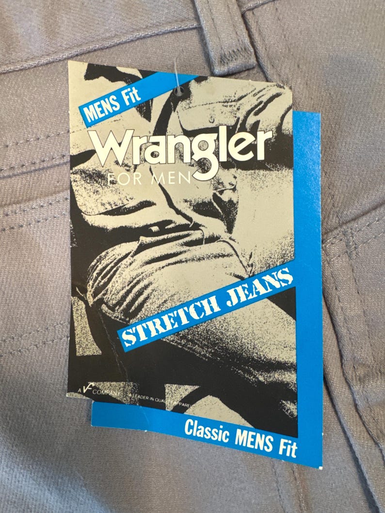 May include: Close-up of light gray Wrangler jeans with a tag. The tag is blue and white with the words "MENS Fit", "Wrangler FOR MEN", and "STRETCH JEANS". The tag also says "Classic MENS Fit".