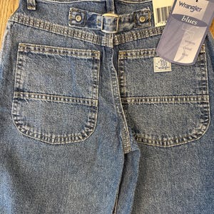 May include: Close-up of the back of blue denim Wrangler jeans. The jeans have two back pockets, a buckle back, and a tag that reads "Wrangler blues western casual wear". The jeans are size 10 regular.