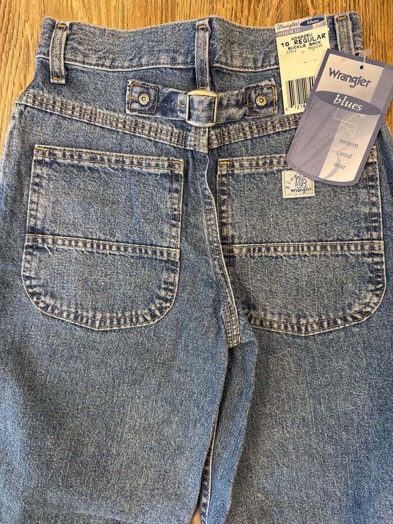 May include: Close-up of the back of blue denim Wrangler jeans. The jeans have two back pockets, a buckle back, and a tag that reads "Wrangler blues western casual wear". The jeans are size 10 regular.