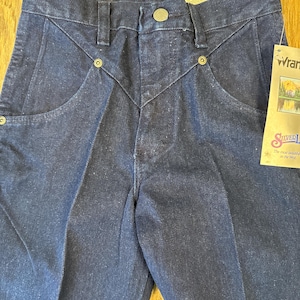 May include: Close-up of dark blue denim jeans with a button closure and decorative stitching. The jeans have a classic five-pocket design and a tag that reads "Wrangler Silver Edition". The jeans are new with tags.