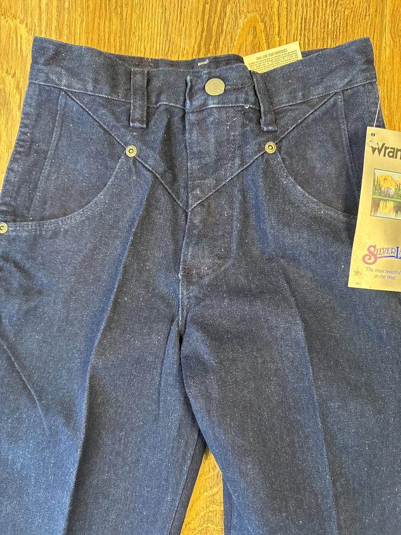 May include: Close-up of dark blue denim jeans with a button closure and decorative stitching. The jeans have a classic five-pocket design and a tag that reads "Wrangler Silver Edition". The jeans are new with tags.