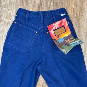 May include: A pair of dark blue denim jeans with a relaxed fit. The jeans have a brown leather patch with the Wrangler logo on the back pocket. The jeans have a tag attached that says "Relaxed Fit Casual Comfort That Stays True To You" and "Wrangler".