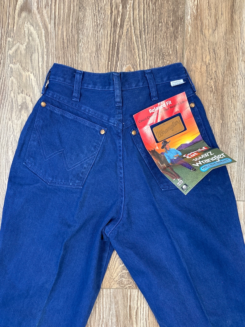 May include: A pair of dark blue denim jeans with a relaxed fit. The jeans have a brown leather patch with the Wrangler logo on the back pocket. The jeans have a tag attached that says "Relaxed Fit Casual Comfort That Stays True To You" and "Wrangler".