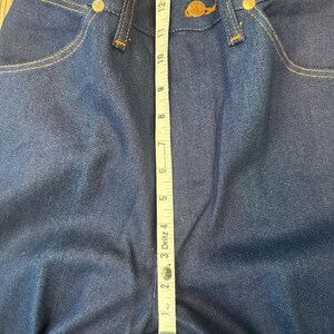 May include: Close-up of dark blue denim jeans with a measuring tape laid across the front. The tape shows a measurement of approximately 15 inches. The jeans have gold-colored rivets and stitching.