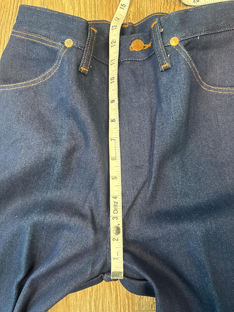 May include: Close-up of dark blue denim jeans with a measuring tape laid across the front. The tape shows a measurement of approximately 15 inches. The jeans have gold-colored rivets and stitching.