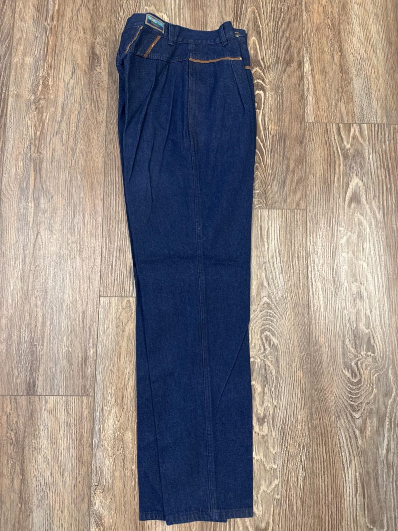 Puede incluir: Un par de pantalones vaqueros de color azul oscuro con ribete de cuero marr&oacute;n alrededor de los bolsillos y la cintura.