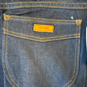 May include: A close-up of a dark blue denim back pocket with orange stitching and a brown leather Wrangler label.