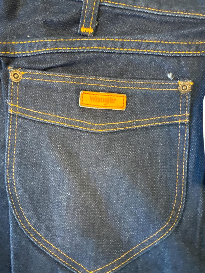 May include: A close-up of a dark blue denim back pocket with orange stitching and a brown leather Wrangler label.