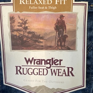 May include: A brown and tan label with the text "Relaxed Fit Fuller Seat & Thigh" and "Wrangler RUGGED WEAR Geared For The Outdoors". The label features a vintage illustration of a man with a dog walking through a mountain landscape.