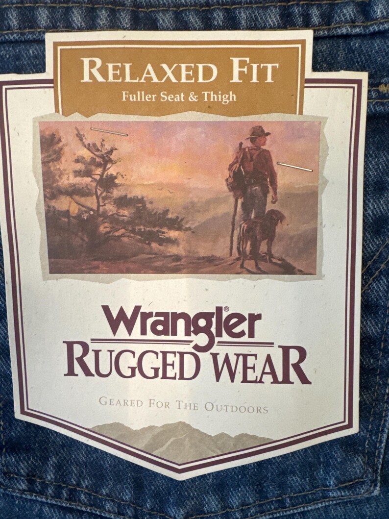 May include: A brown and tan label with the text "Relaxed Fit Fuller Seat & Thigh" and "Wrangler RUGGED WEAR Geared For The Outdoors". The label features a vintage illustration of a man with a dog walking through a mountain landscape.