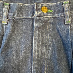 May include: Close-up of blue denim jeans with a button closure. The jeans have green stitching accents on the belt loops and a gold button. The denim fabric has a faded appearance.