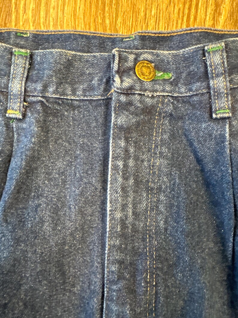 May include: Close-up of blue denim jeans with a button closure. The jeans have green stitching accents on the belt loops and a gold button. The denim fabric has a faded appearance.