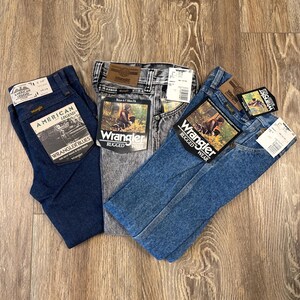 Puede incluir: Tres pares de pantalones vaqueros azules, uno azul oscuro, uno azul claro y uno gris. Los vaqueros est&aacute;n todos doblados y tienen etiquetas de Wrangler adjuntas.