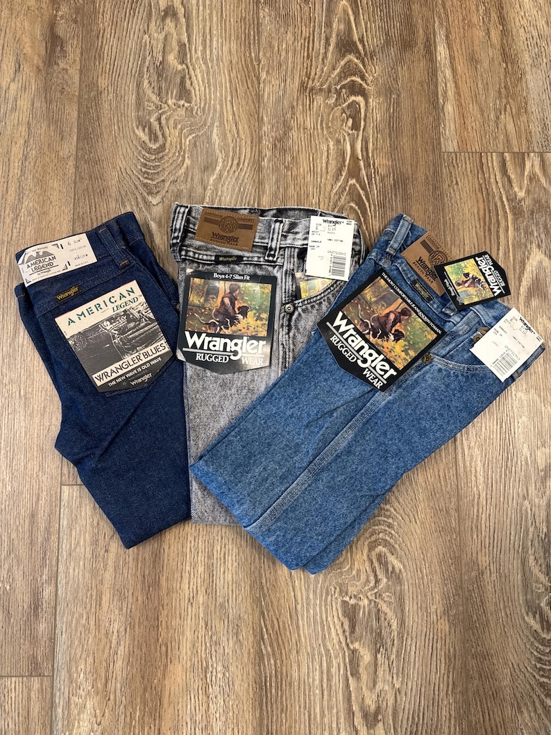 Puede incluir: Tres pares de pantalones vaqueros azules, uno azul oscuro, uno azul claro y uno gris. Los vaqueros est&aacute;n todos doblados y tienen etiquetas de Wrangler adjuntas.