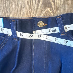K&ouml;nnte beinhalten: Nahaufnahme einer dunkelblauen Jeans mit braunem Lederbund. Ein Ma&szlig;band ist um den Bund gewickelt und zeigt Ma&szlig;e von 25 bis 32 Zoll.