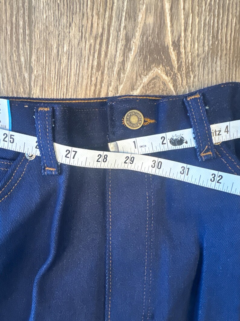 K&ouml;nnte beinhalten: Nahaufnahme einer dunkelblauen Jeans mit braunem Lederbund. Ein Ma&szlig;band ist um den Bund gewickelt und zeigt Ma&szlig;e von 25 bis 32 Zoll.