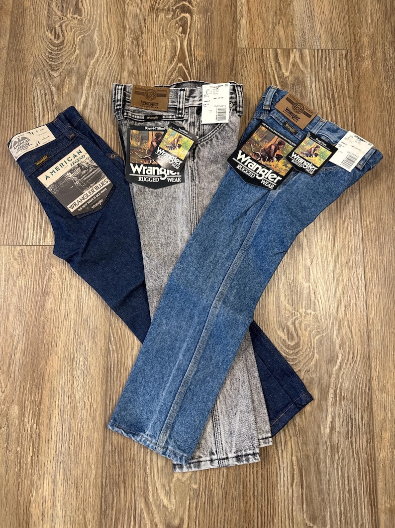 Puede incluir: Tres pares de pantalones vaqueros de mezclilla azul, un par es un lavado azul oscuro, un par es un lavado azul claro y un par es un lavado gris claro con un patr&oacute;n moteado. Los tres pares tienen una etiqueta Wrangler y las palabras "Rugged Wear" en la etiqueta.