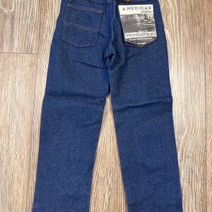 Puede incluir: Un par de pantalones vaqueros de mezclilla azul oscuro con una etiqueta Wrangler American Legend Wrangler Blues adjunta. La etiqueta dice "The new wave is old wave".
