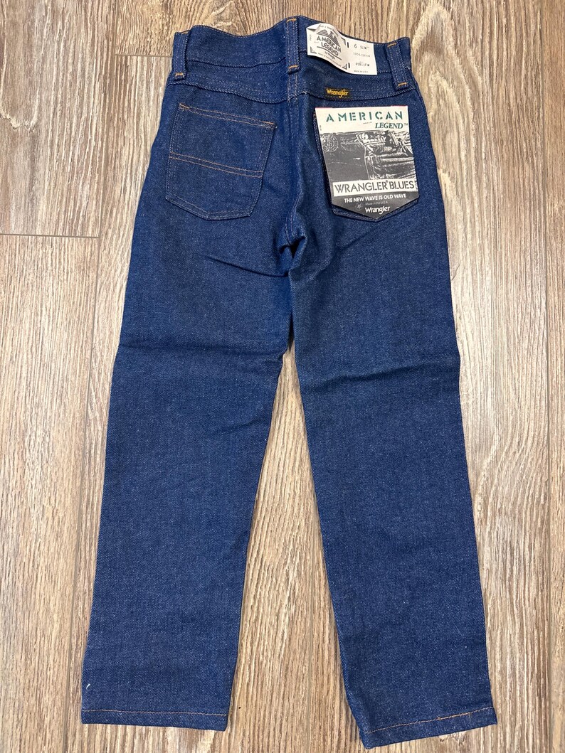 Puede incluir: Un par de pantalones vaqueros de mezclilla azul oscuro con una etiqueta Wrangler American Legend Wrangler Blues adjunta. La etiqueta dice "The new wave is old wave".