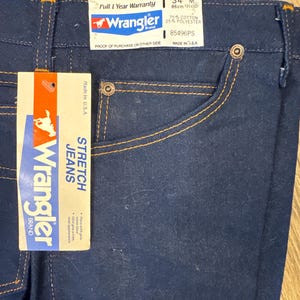 May include: Dark blue Wrangler stretch jeans with orange stitching and a tag. The tag reads "Wrangler Brand STRETCH JEANS" and includes the size 34M, which is equivalent to a 31/12 waist size. The jeans are made in the USA.