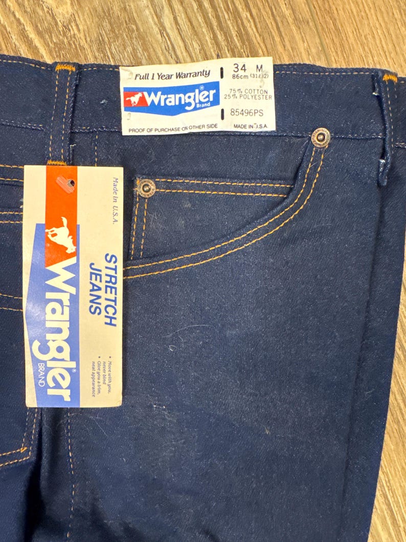 May include: Dark blue Wrangler stretch jeans with orange stitching and a tag. The tag reads "Wrangler Brand STRETCH JEANS" and includes the size 34M, which is equivalent to a 31/12 waist size. The jeans are made in the USA.
