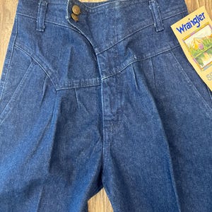 Puede incluir: Primer plano de unos vaqueros Wrangler de mezclilla azul vintage. Los vaqueros presentan una cintura alta, pliegues y un cierre de bot&oacute;n &uacute;nico. Una etiqueta Wrangler es visible en el lado derecho. La mezclilla tiene un aspecto cl&aacute;sico, ligeramente descolorido.