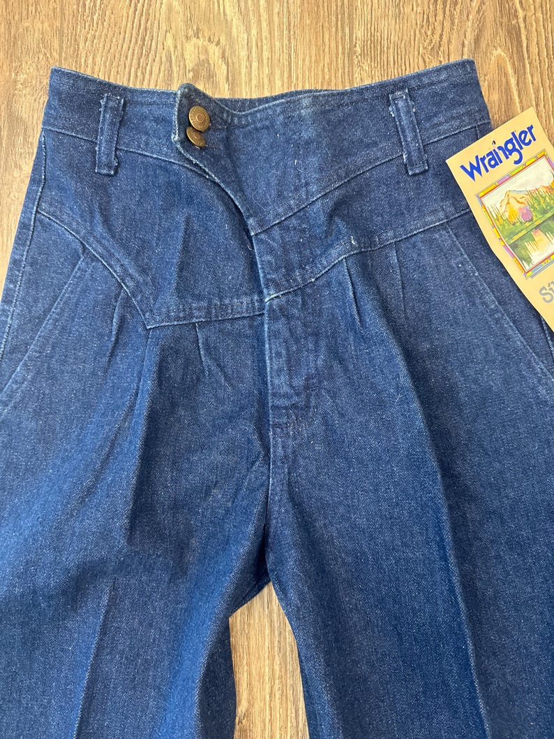 Puede incluir: Primer plano de unos vaqueros Wrangler de mezclilla azul vintage. Los vaqueros presentan una cintura alta, pliegues y un cierre de bot&oacute;n &uacute;nico. Una etiqueta Wrangler es visible en el lado derecho. La mezclilla tiene un aspecto cl&aacute;sico, ligeramente descolorido.