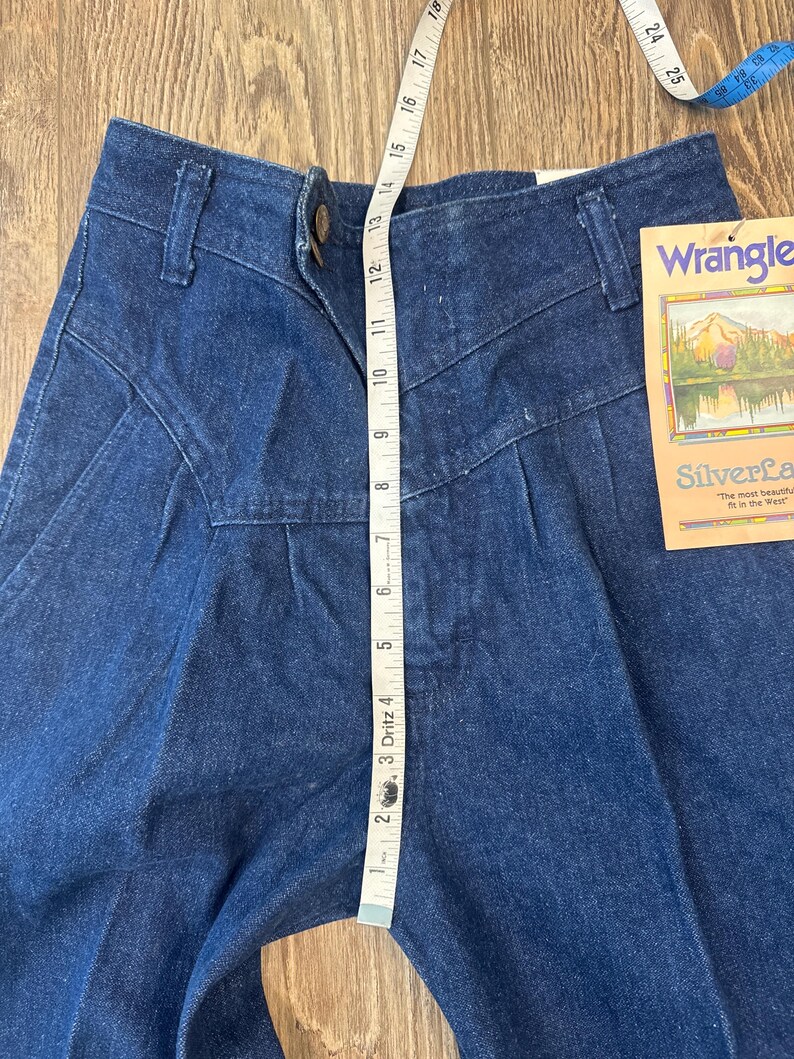 Puede incluir: Pantalones vaqueros azul oscuro con cintura alta y cierre de bot&oacute;n. Una cinta m&eacute;trica muestra una longitud de aproximadamente 33 cm. Los pantalones tienen una etiqueta que dice "Wrangler Silverlake".
