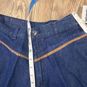 Puede incluir: Un par de pantalones vaqueros de mezclilla azul oscuro con ribetes de cuero marr&oacute;n. Los pantalones tienen cierre de bot&oacute;n y una cintura con una cinta m&eacute;trica que muestra un tama&ntilde;o de 13 pulgadas.