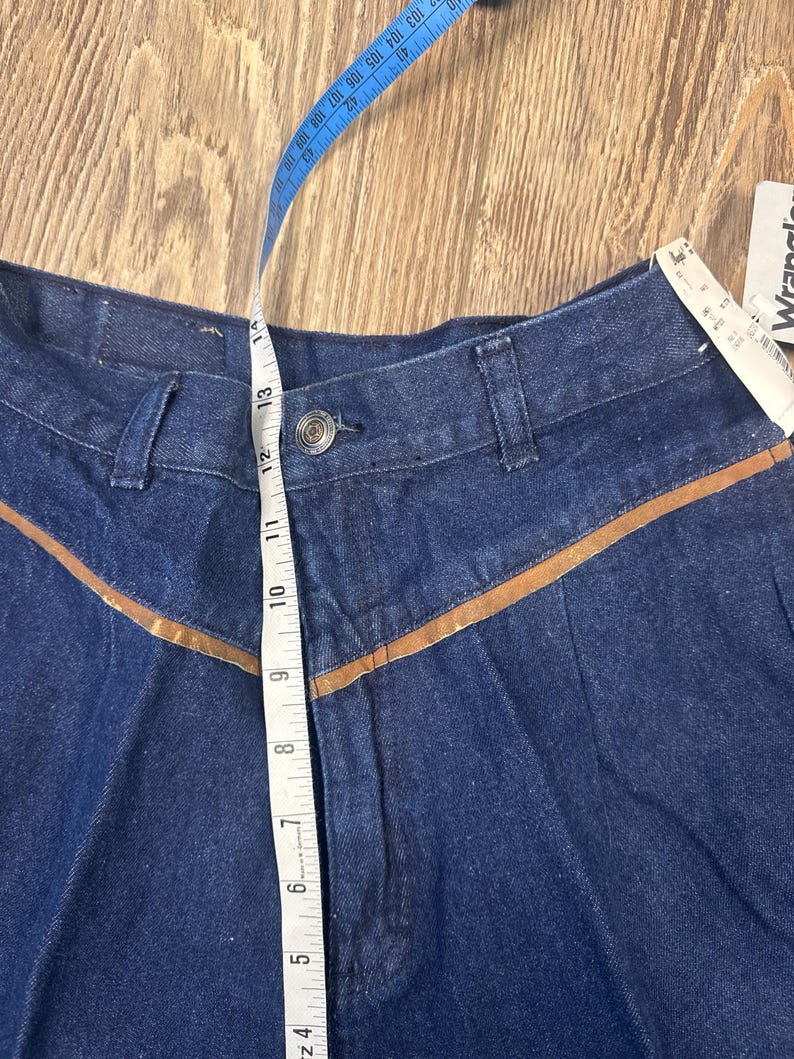 Puede incluir: Un par de pantalones vaqueros de mezclilla azul oscuro con ribetes de cuero marr&oacute;n. Los pantalones tienen cierre de bot&oacute;n y una cintura con una cinta m&eacute;trica que muestra un tama&ntilde;o de 13 pulgadas.