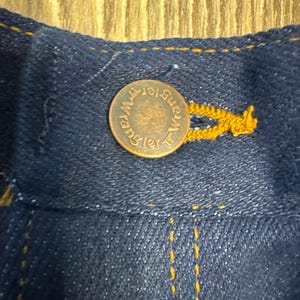 May include: Close-up of a dark blue denim fabric with a copper-colored button. The button is embossed with the word "Wrangler" in a circular pattern. Yellow stitching is visible along the seams, highlighting the button and fabric.