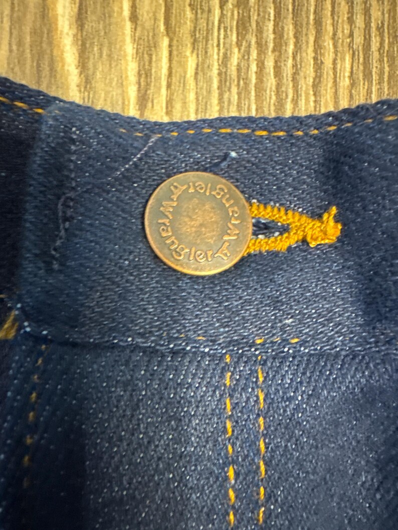 May include: Close-up of a dark blue denim fabric with a copper-colored button. The button is embossed with the word "Wrangler" in a circular pattern. Yellow stitching is visible along the seams, highlighting the button and fabric.