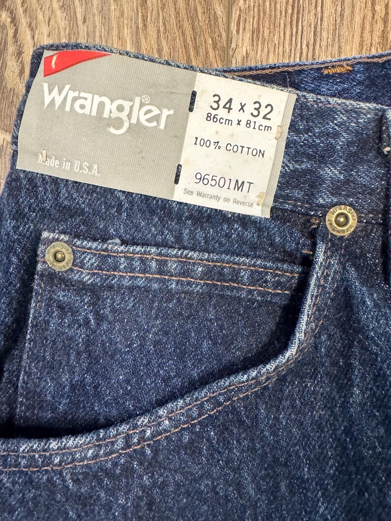 May include: Close-up of dark blue Wrangler jeans. The label indicates a size of 34 x 32 inches (86cm x 81cm) and states the jeans are 100% cotton. The tag also says "Made in U.S.A."