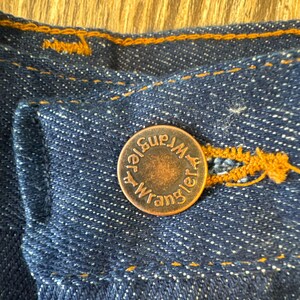May include: Close-up of a copper-colored button with the text "Wrangler" and "Blue Bell" on it. The button is sewn onto a dark blue denim fabric.