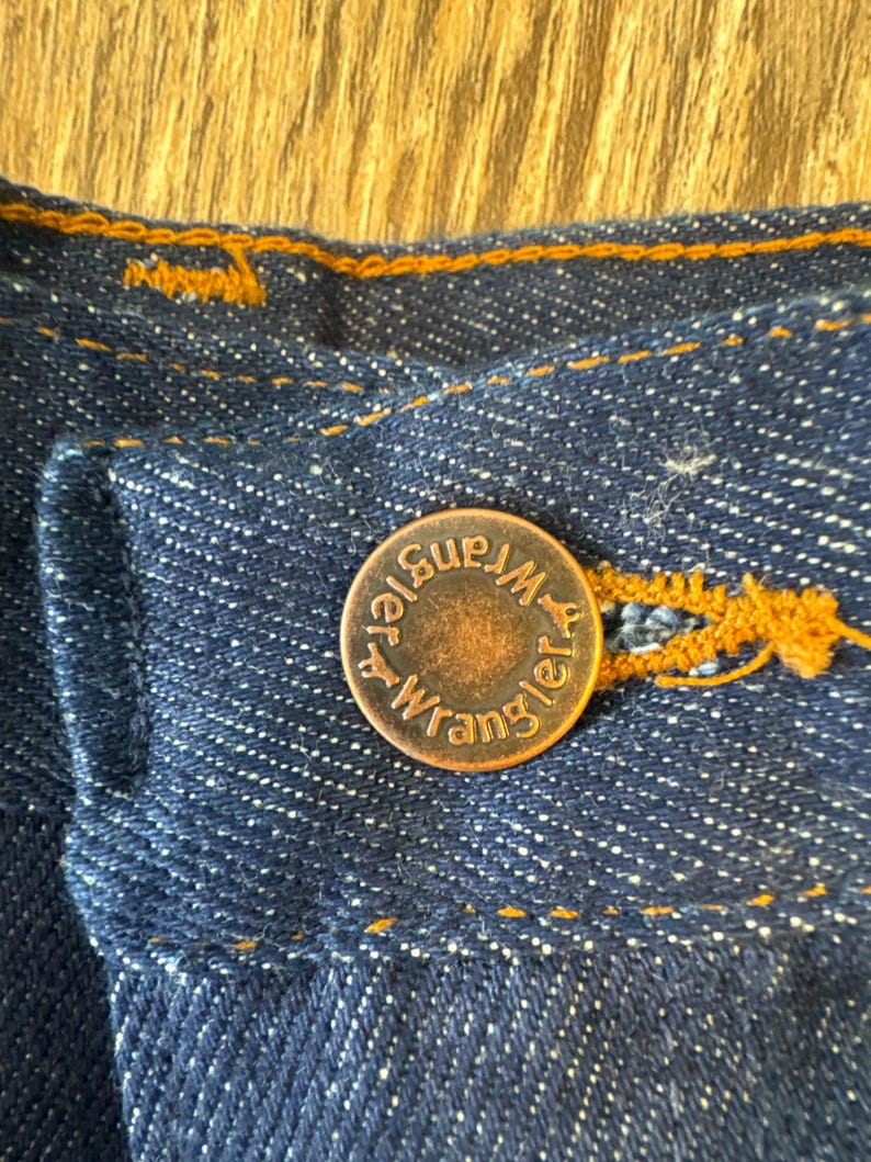May include: Close-up of a copper-colored button with the text "Wrangler" and "Blue Bell" on it. The button is sewn onto a dark blue denim fabric.