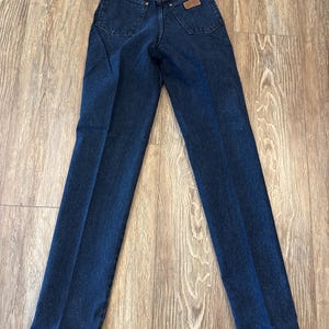 May include: Dark blue denim jeans with a classic five-pocket design. The jeans have a straight leg cut and a brown leather patch on the back pocket. A red tag with the brand name is attached.