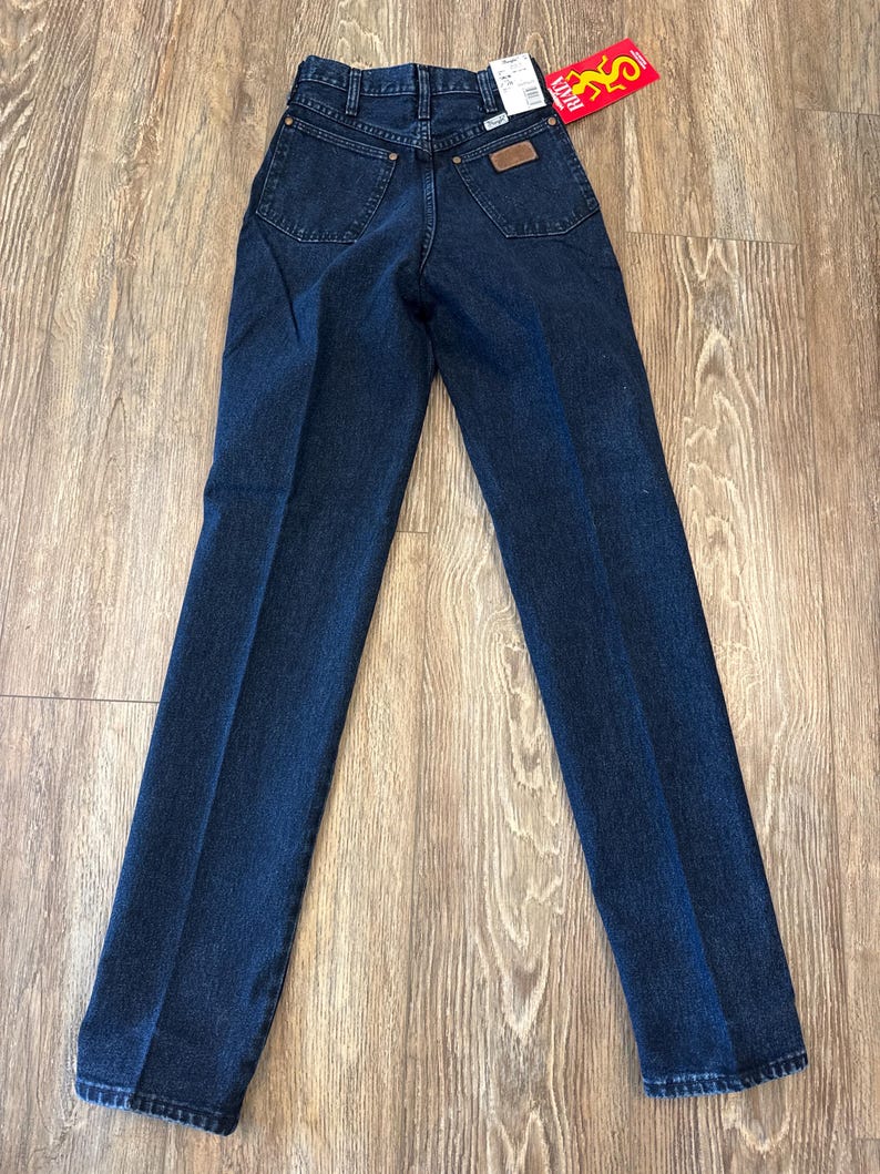 May include: Dark blue denim jeans with a classic five-pocket design. The jeans have a straight leg cut and a brown leather patch on the back pocket. A red tag with the brand name is attached.