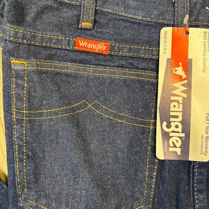 May include: Close-up of dark blue denim jeans with orange stitching. The back pocket features a classic design. A red Wrangler brand tag is visible. A paper tag with the Wrangler logo and text is attached.
